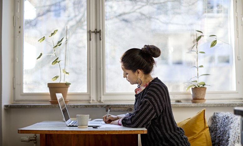 Kvinna sitter vid ett bord och skriver på ett papper med datorn framför sig. Ljuset strålar in från ett stort fönster bakom henne