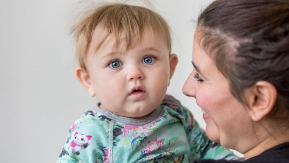 Kvinna med mörkt hår håller i en bebis med grön tröja med lila blommor på.